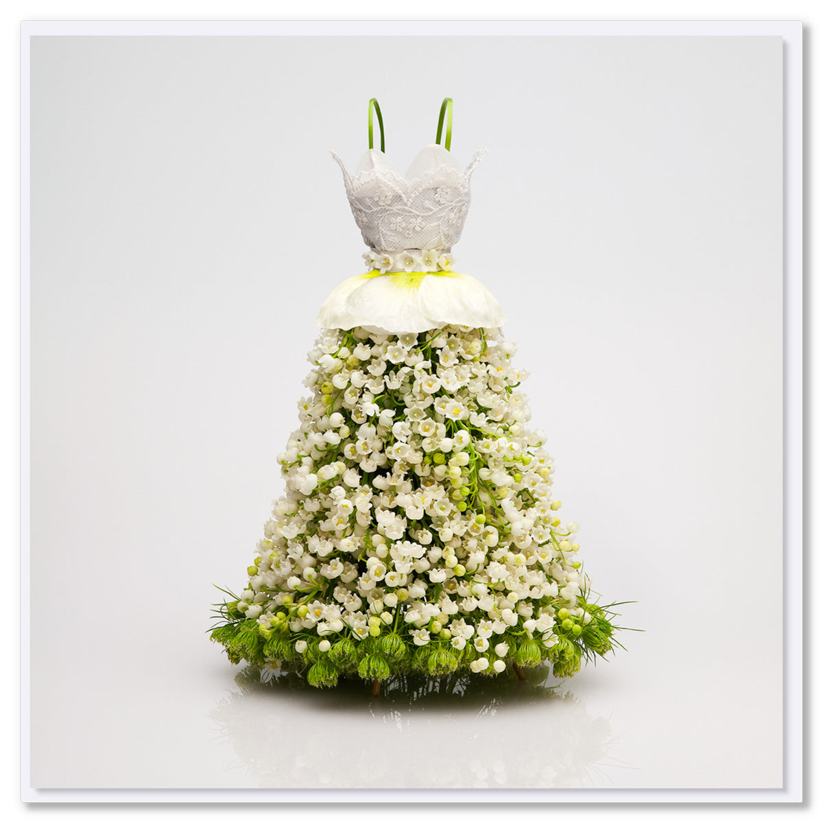 Floral dress made of white blossoms with intricate lace bodice, full skirt, green calla lily straps, and green flowers at base against white background.