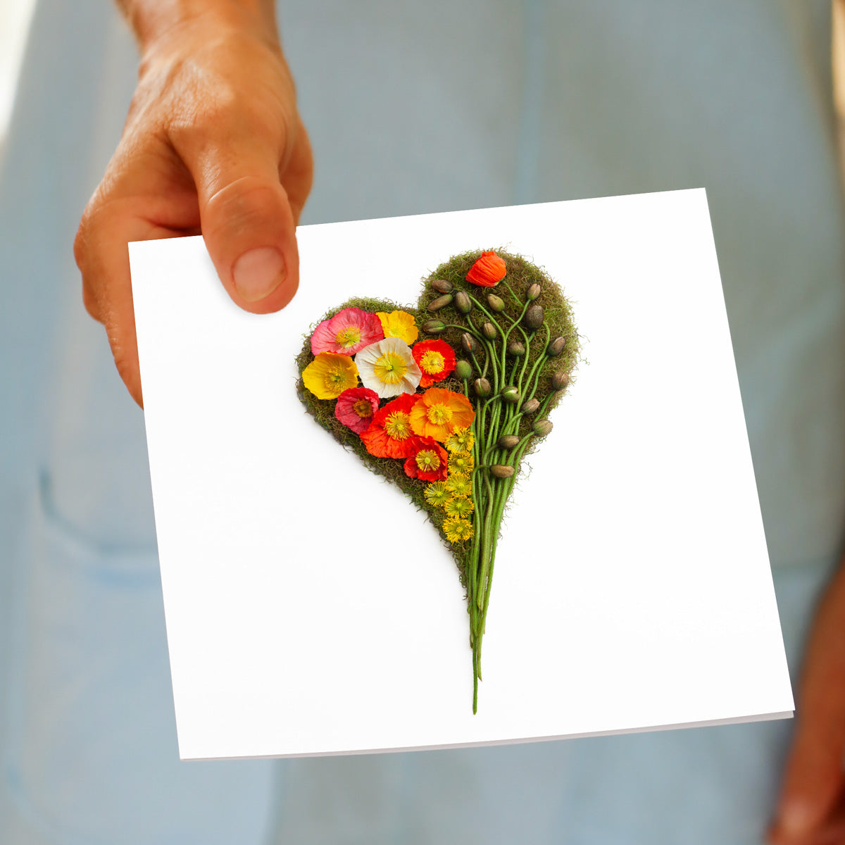 A held by hand poppy heart greeting card from Golly Gosh Creations. A handcrafted moss heart adorned with vibrant wild poppies and buds. Size 142 x 142 mm.