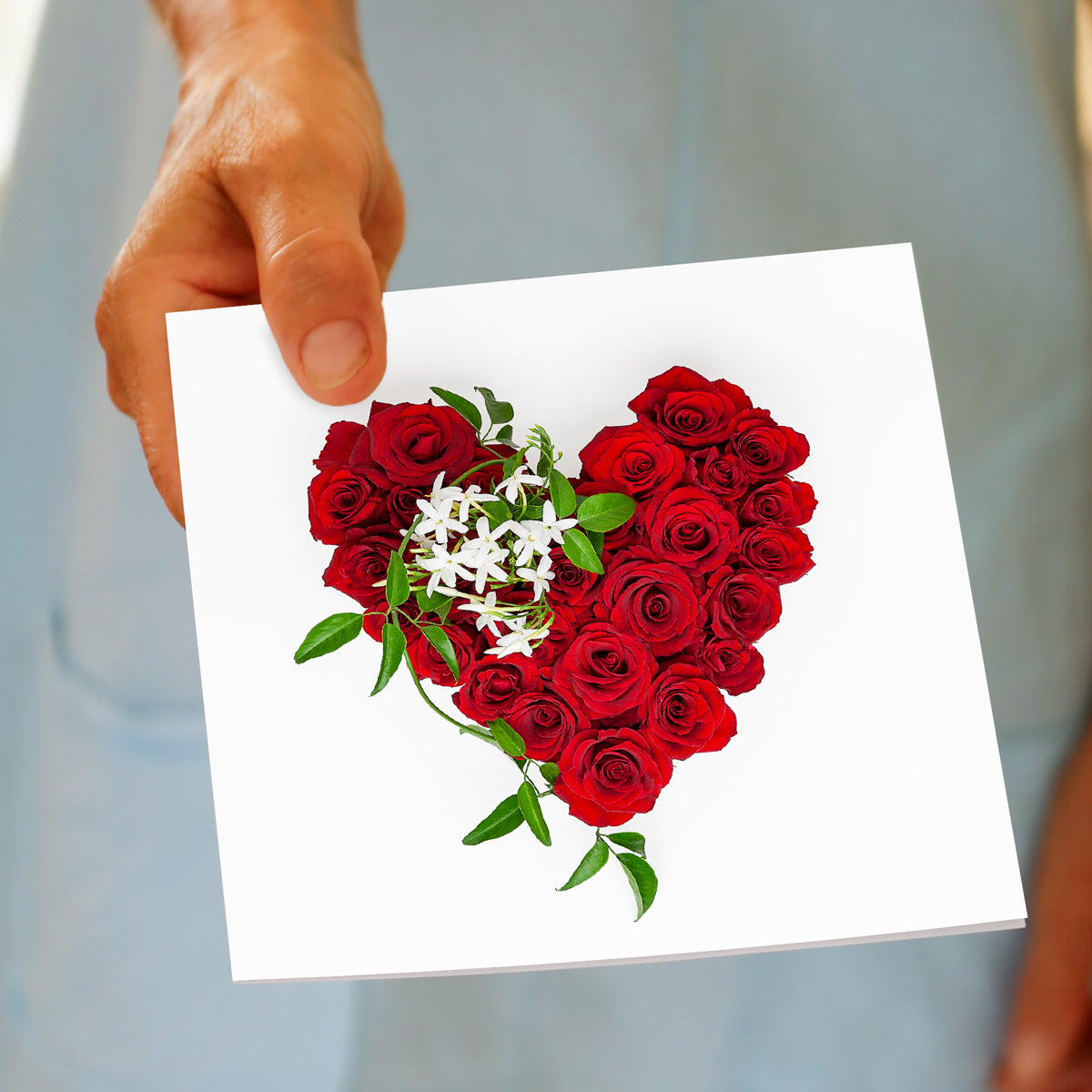 A red rose heart greeting card from Golly Gosh Creations. This generous square card is being held by hand. Created from fresh vibrant red roses.