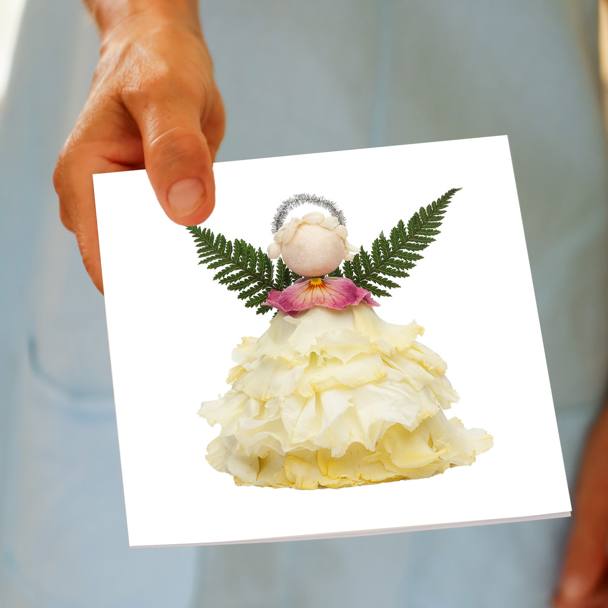 A hand holds the Christmas Cream Angel greeting card, featuring a cream rose flower angel with fern wings, a pink flower collar, and a silver halo, made from fresh flowers by Golly Gosh Creations.
