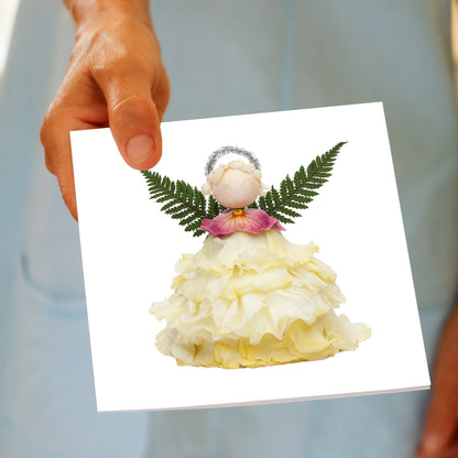 A hand holds the Christmas Cream Angel greeting card, featuring a cream rose flower angel with fern wings, a pink flower collar, and a silver halo, made from fresh flowers by Golly Gosh Creations.