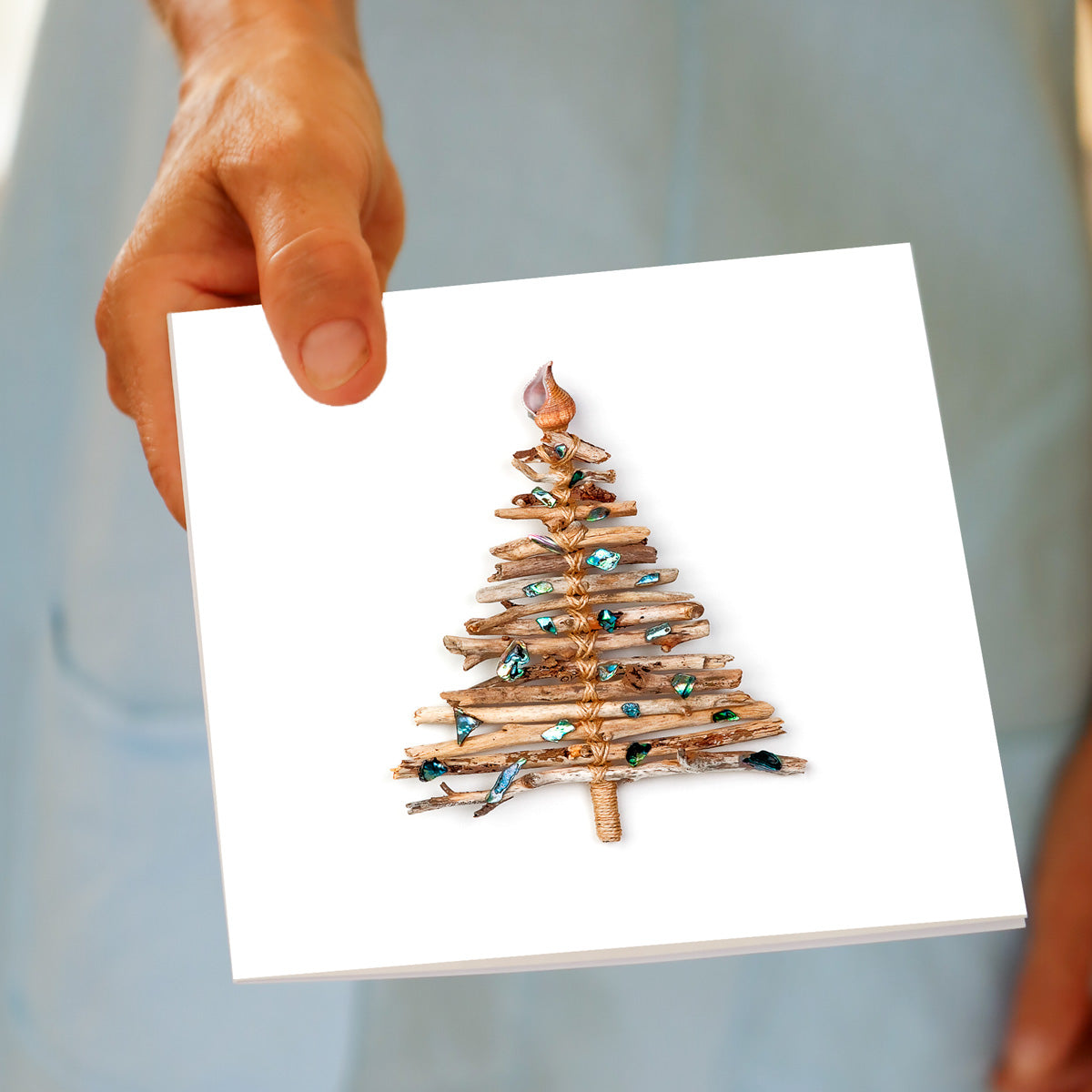 A hand holds the Christmas Driftwood Tree greeting card, featuring driftwood bundled into a tree shape with paua shell pieces and a spiral shell topper, made by Golly Gosh Creations.