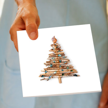 A hand holds the Christmas Driftwood Tree greeting card, featuring driftwood bundled into a tree shape with paua shell pieces and a spiral shell topper, made by Golly Gosh Creations.