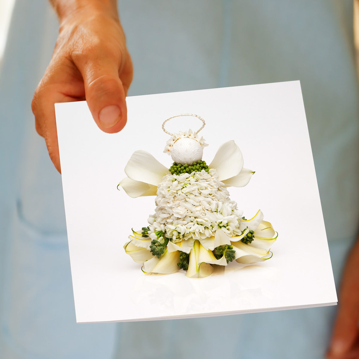 A hand holds the Christmas Lily Angel greeting card, featuring a white lily flower angel with a halo and a collar of green buds, made from fresh flowers by Golly Gosh Creations.