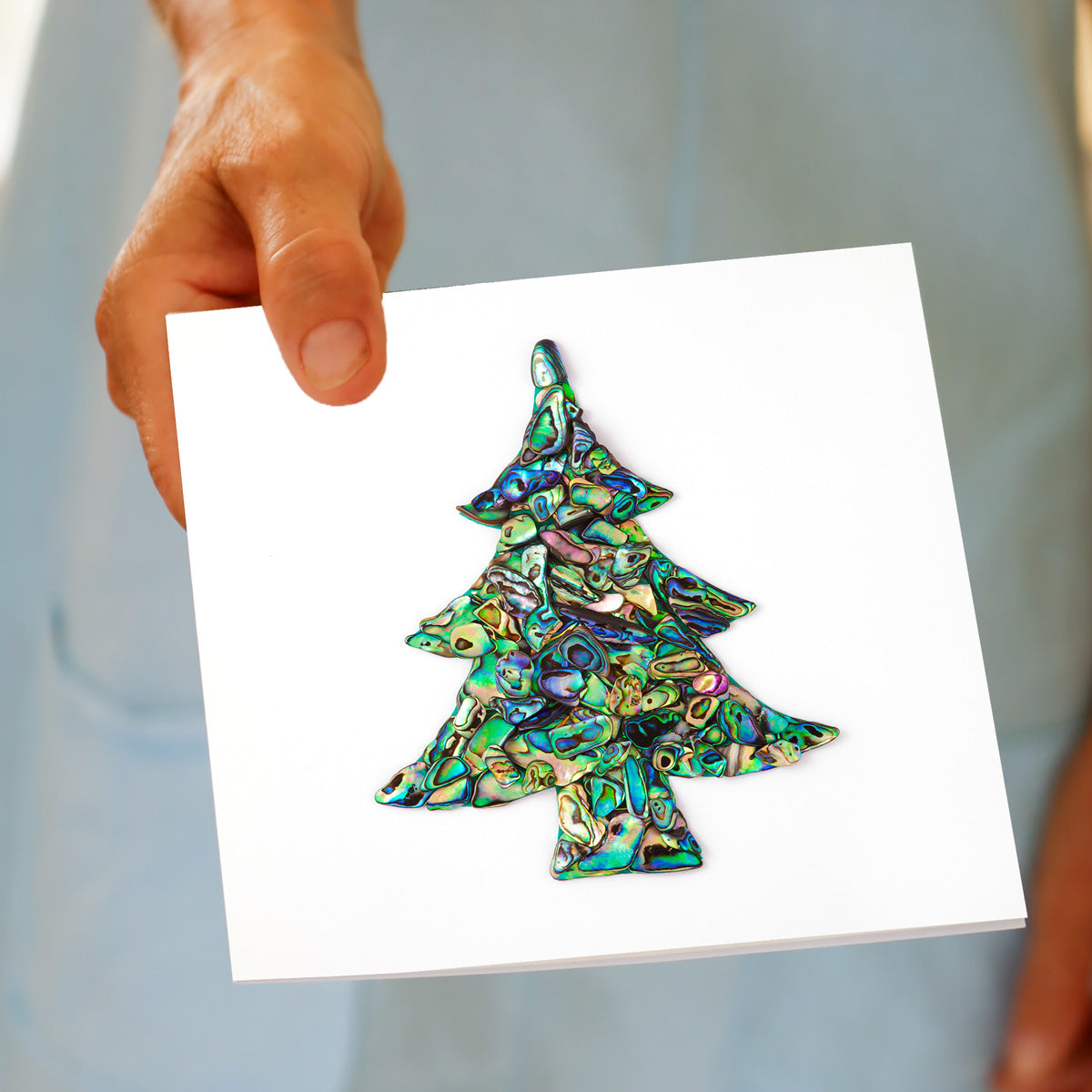 A hand holds the Christmas Paua Tree greeting card, featuring a Christmas tree shape made from iridescent paua shell pieces in green, blue, and purple, made by Golly Gosh Creations.