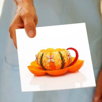 A pumpkin cup greeting card by Golly Gosh Creations held by hand. Size 142 x 142 mm. Created from bright pumpkin, fruits and seeds with a red chilli handle.