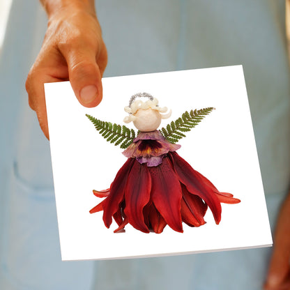 A hand holds the Christmas Red Angel greeting card, featuring a red lily flower angel with fern wings and a silver halo, made from fresh flowers by Golly Gosh Creations.