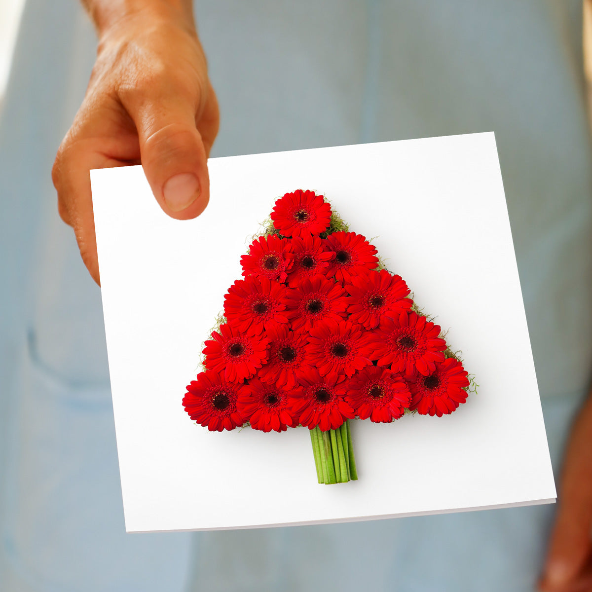 A hand holds the Christmas Gerbera Tree greeting card, featuring red gerbera daisies built into a tree shape with green moss and bundled stems, made by Golly Gosh Creations.