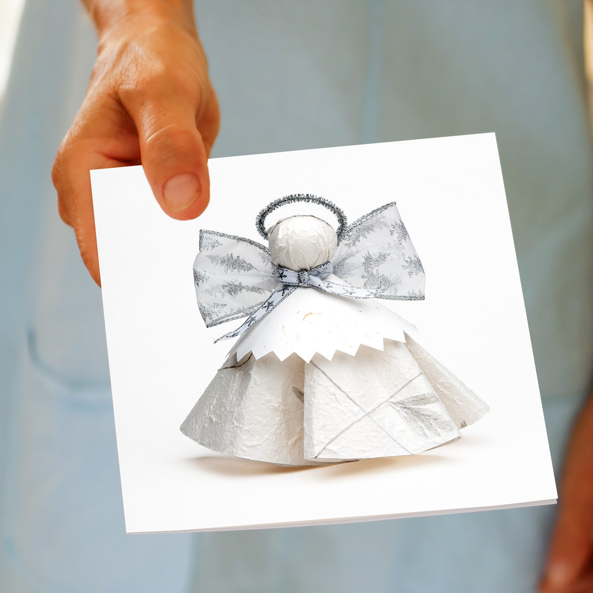 A hand holds the Christmas Silver Angel greeting card, featuring a white paper angel with a silver and white ribbon bow and a fine halo, made by Golly Gosh Creations.