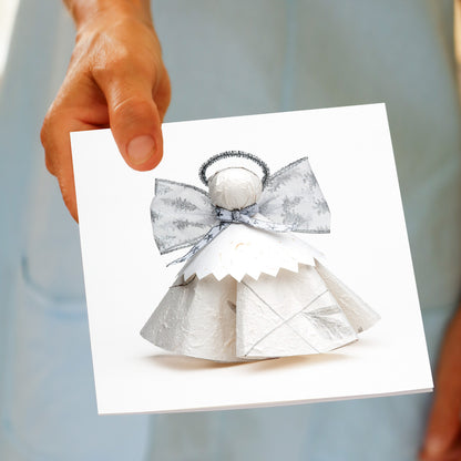 A hand holds the Christmas Silver Angel greeting card, featuring a white paper angel with a silver and white ribbon bow and a fine halo, made by Golly Gosh Creations.