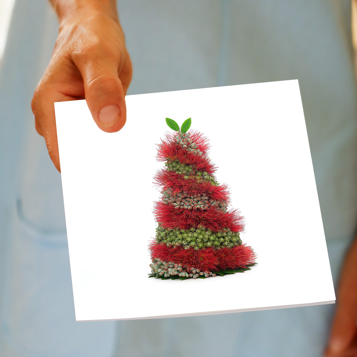 A hand holds the Christmas Spiral Pohutukawa Tree greeting card, featuring red pohutukawa flowers and buds spiralling into a tree shape, made by Golly Gosh Creations.