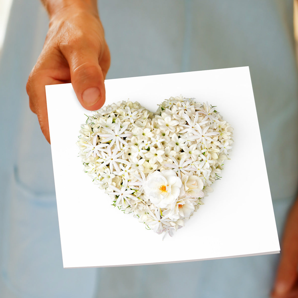 A stephanotis heart greeting card from Golly Gosh Creations, being held by hand. Created from fresh white stephanotis blossoms. Size 142 x 142 mm.